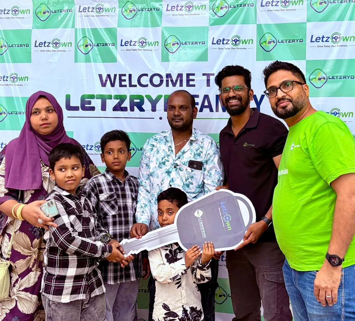 A driver and his family holding an oversized LetzOwn key in front of a Welcome to the LetzRyd Family backdrop